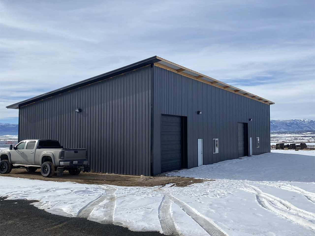 A gray steel building with large overhead doors, set in a snowy landscape with a parked pickup truck nearby.